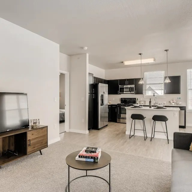 Contemporary Apartment Interior A modern apartment living area featuring a gray sofa, a small round coffee table, a TV unit with a flat-screen television, and bar stools at a kitchen island in the background.