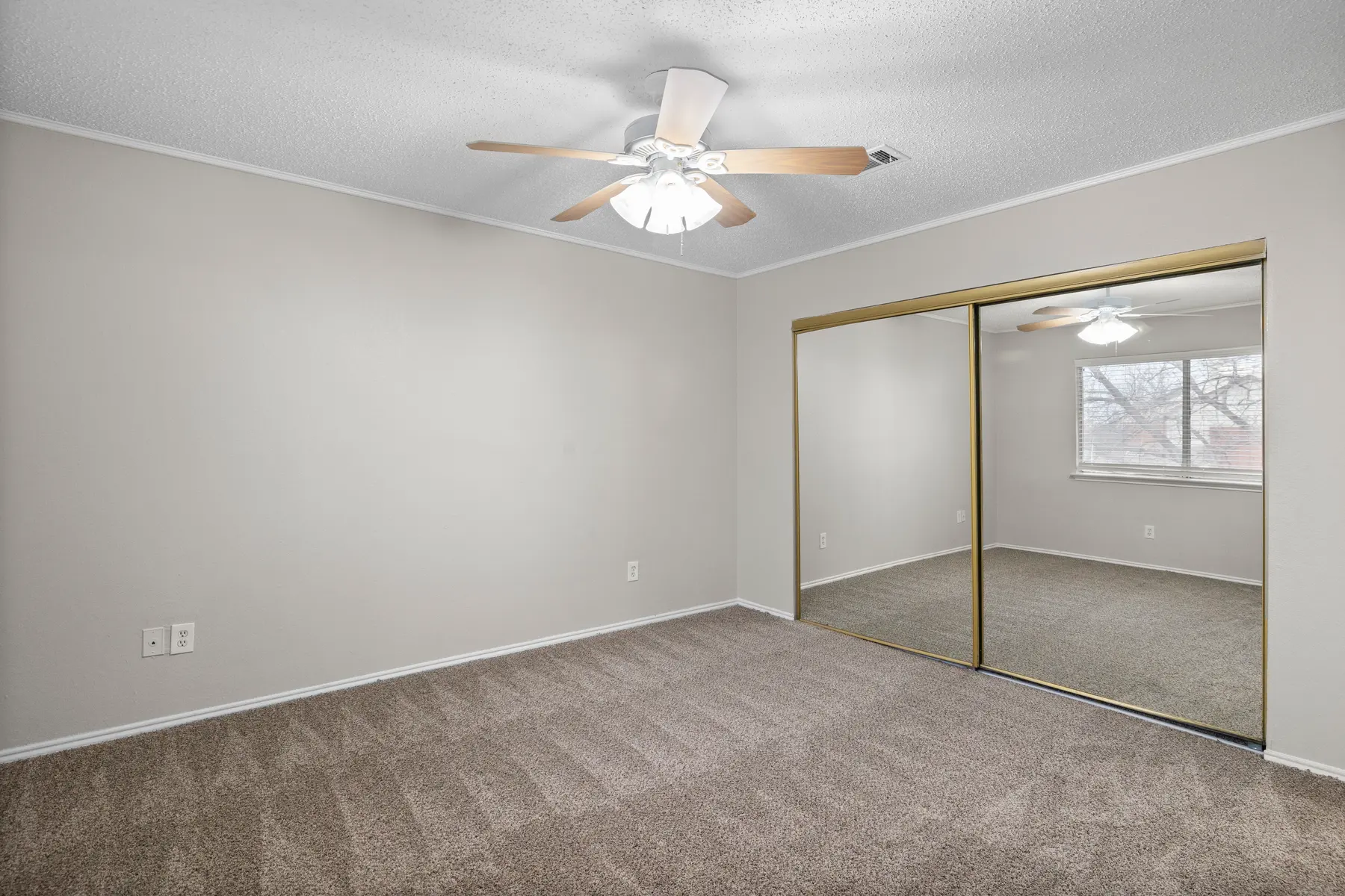 A spacious, empty bedroom featuring light gray walls and plush carpet. The room has a ceiling fan with wooden blades and a large mirrored closet on one wall. Natural light enters from a window on the opposite wall.