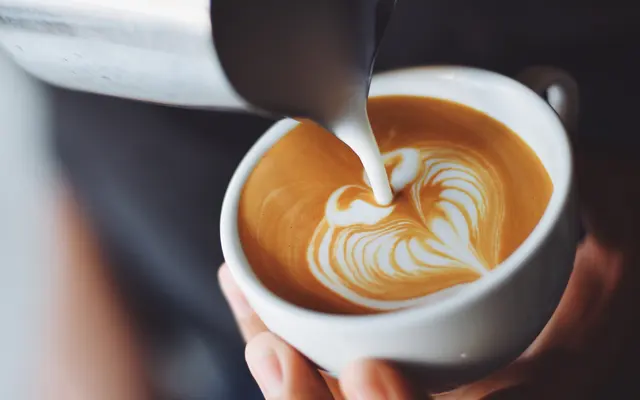 A close-up image of a hand pouring steamed milk from a metal pitcher into a cup of coffee, creating latte art with a heart design.
