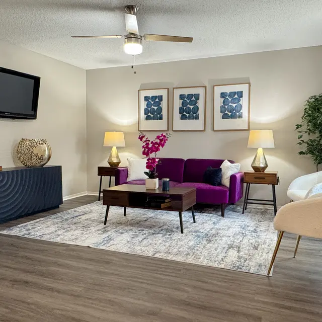 A modern living room featuring a purple sofa, two lamps, a coffee table, and a TV on the wall. There are framed artworks above the sofa and a potted plant in the corner.