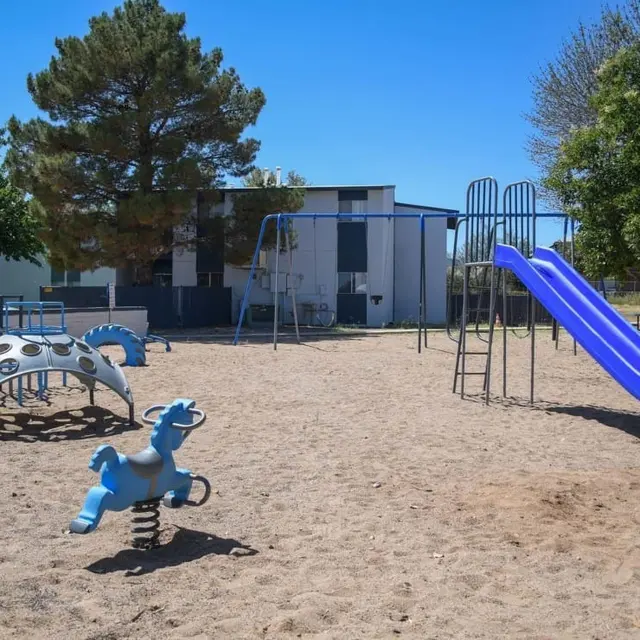 A sunny playground featuring a blue slide, a climbing structure, and a spring rider horse. The ground is covered in sand, surrounded by trees and a residential building in the background.