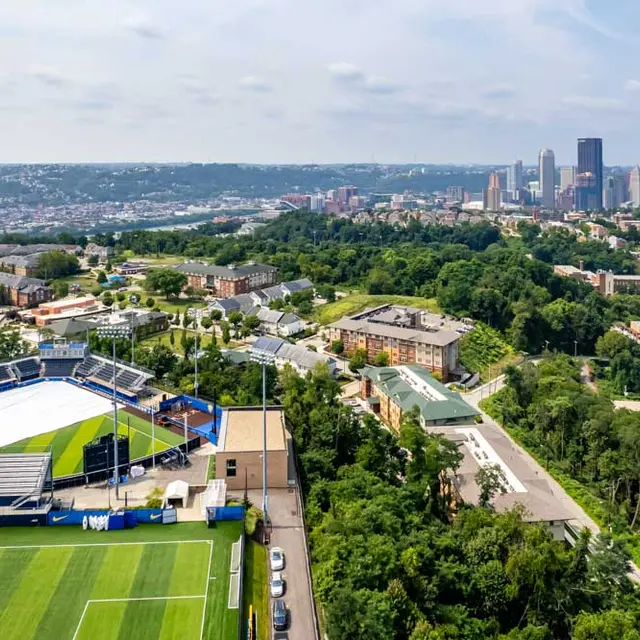 Aerial view of a soccer stadium with green fields surrounded by residential and urban areas, showcasing a city skyline in the background.