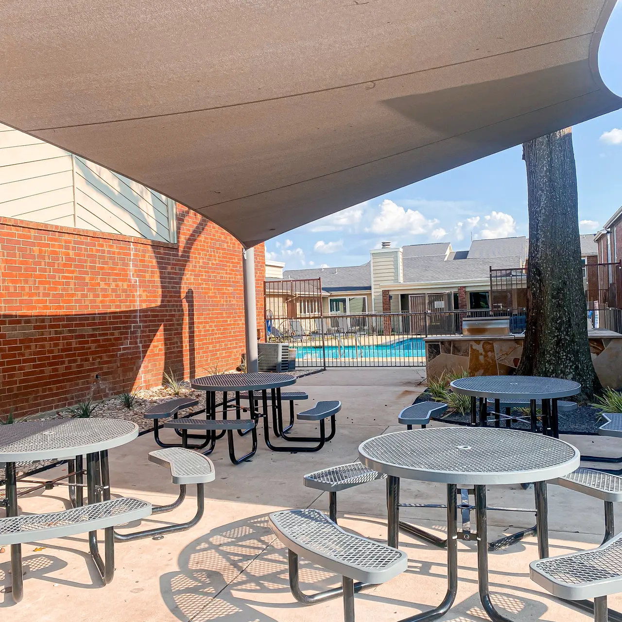 A shaded outdoor dining area with several round tables and chairs, surrounded by a pool and buildings in the background.