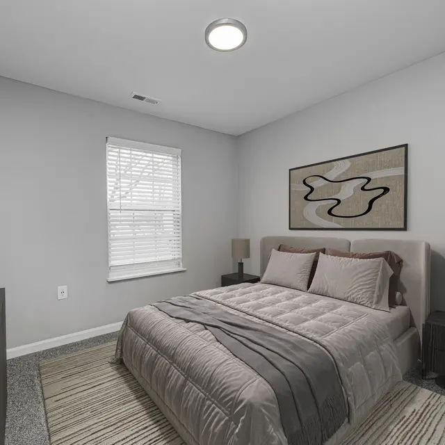A modern bedroom featuring a large bed with light gray bedding, two nightstands with lamps, and a piece of abstract wall art. The room has gray walls and carpet, a window with blinds, and a minimalist decor style.