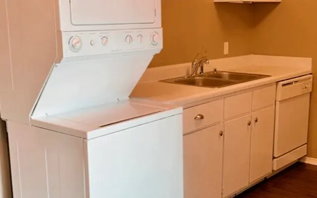 Compact Kitchen with Laundry Unit A compact kitchen area featuring a stacked washer and dryer unit, a sink, and a dishwasher, with light colored cabinets and wooden flooring.