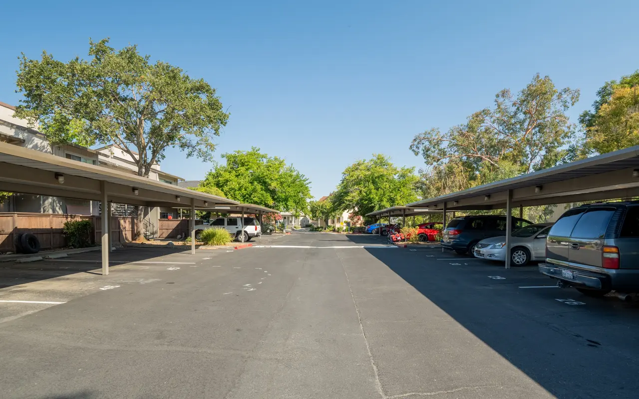 A view of an open parking lot with covered spaces, surrounded by trees and residential buildings nearby under a clear blue sky.