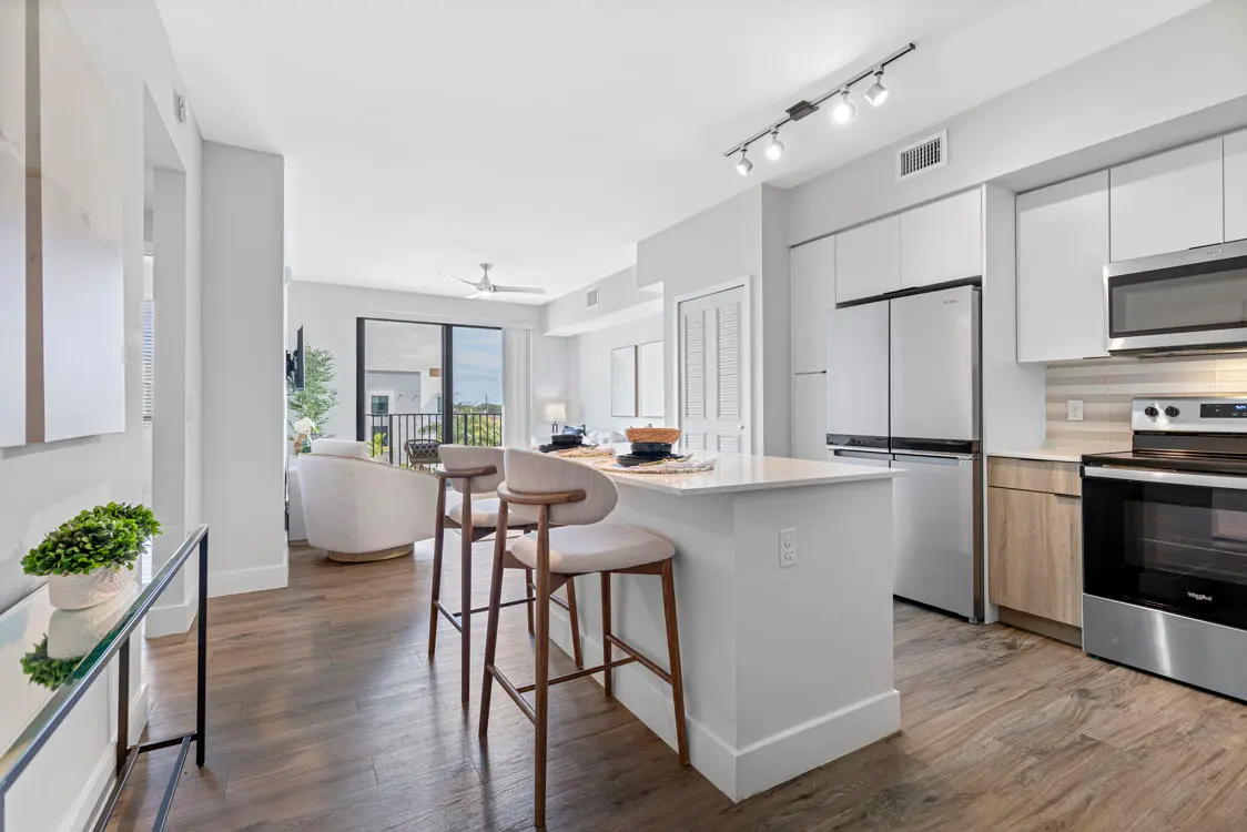 Contemporary Open Kitchen Design A modern kitchen with an open layout featuring a breakfast bar, wood flooring, and large windows offering natural light. There are stools at the bar and a cozy seating area in the background adjacent to a sliding glass door leading to a balcony.