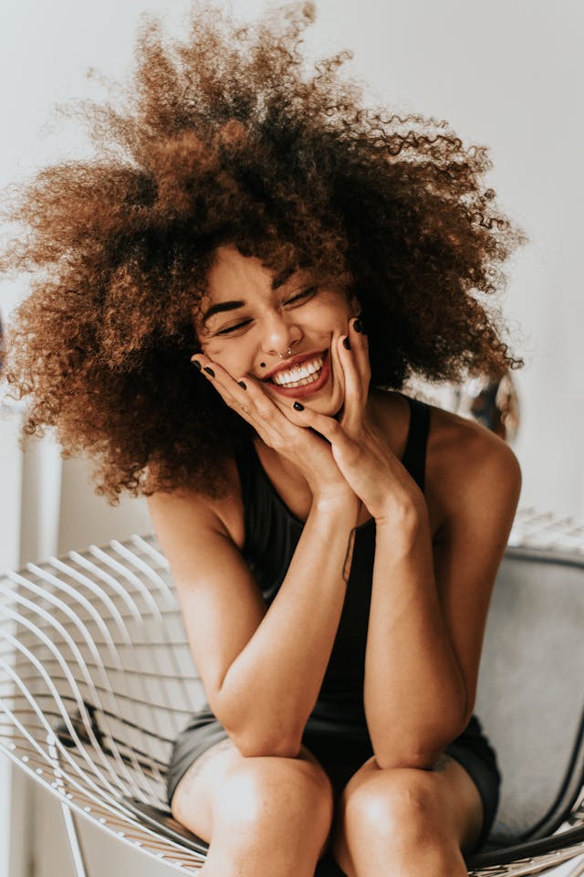 Joyful Woman with Natural Curls A smiling woman with curly, voluminous hair sitting in a modern chair, holding her face with her hands in a joyful expression.