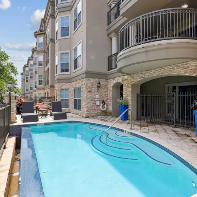 A modern apartment complex featuring a small pool area surrounded by a patio and lounge chairs, with a decorative fence and lush green trees in the background.