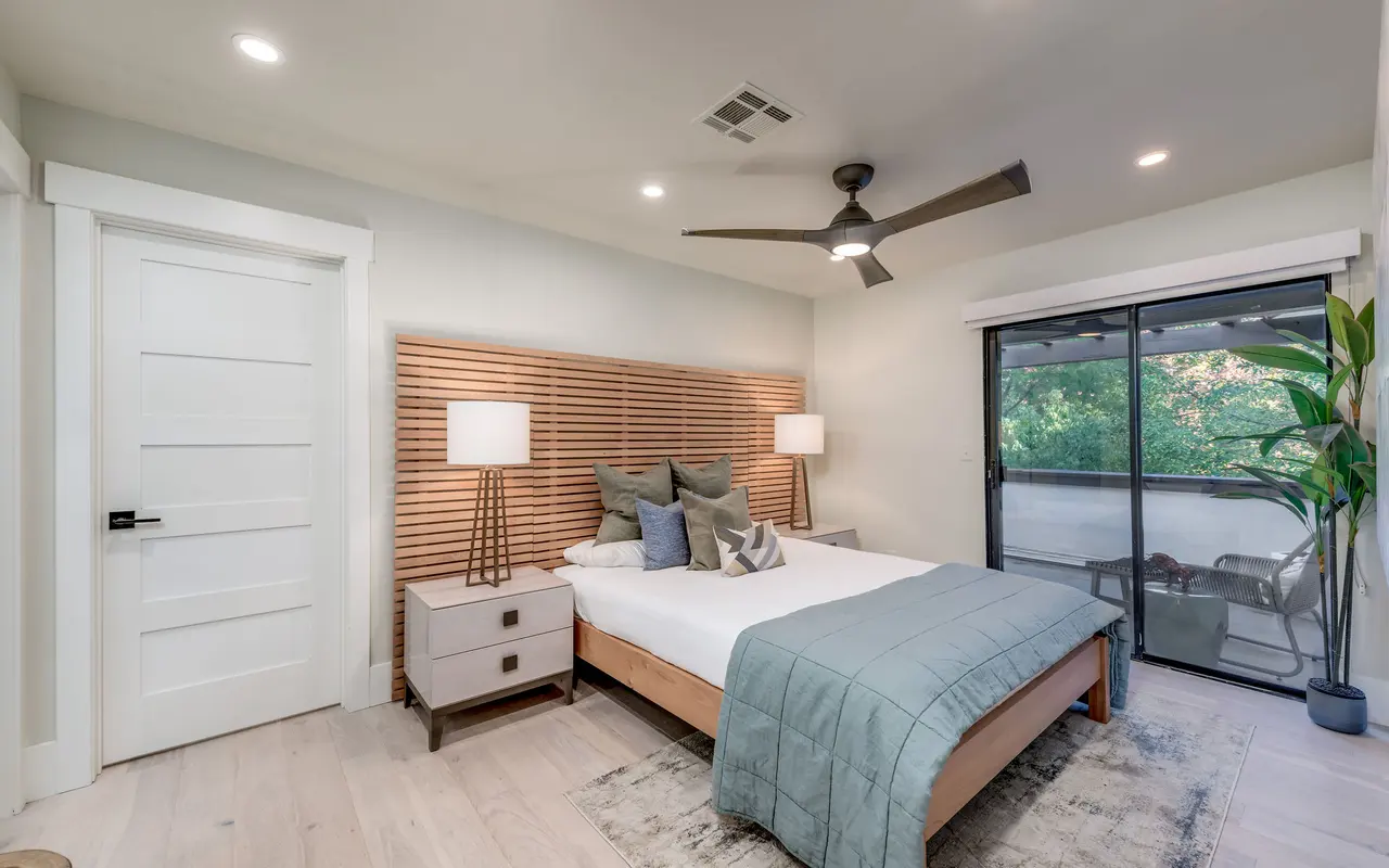 A modern bedroom featuring a bed with a wooden headboard, decorative pillows, two bedside lamps on nightstands, and a large window overlooking greenery.