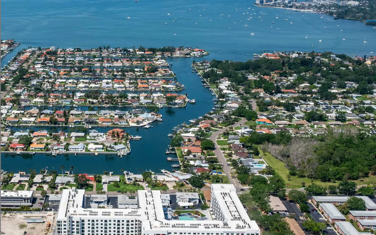 Aerial view of a waterfront residential area with canals and houses, along with a nearby building complex and greenery.