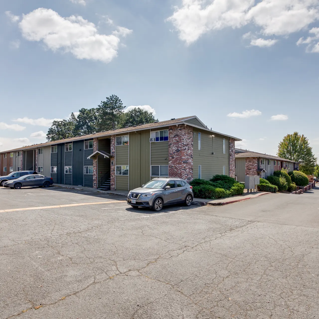 Apartment Complex Exterior View Exterior view of an apartment complex with two-story buildings, surrounded by trees and a parking lot filled with cars.