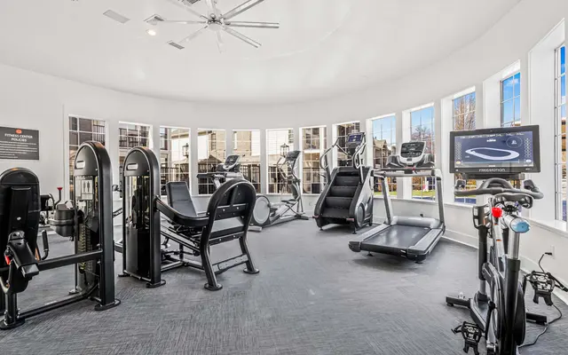 A modern gym with multiple workout machines, including treadmills and weight training equipment, situated in a bright room with large windows.