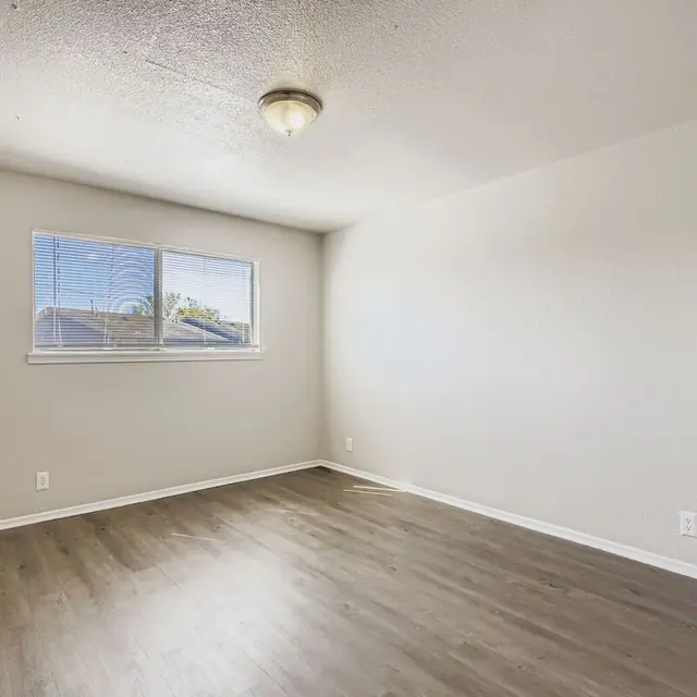 An empty room with light-colored walls and brown wooden flooring. There is a window on one wall allowing natural light to enter, and a ceiling light fixture.