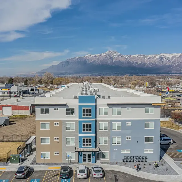 A modern multi-story apartment building with a light blue and beige exterior. In the background, there are snow-capped mountains, and the surrounding area includes other buildings and open land.