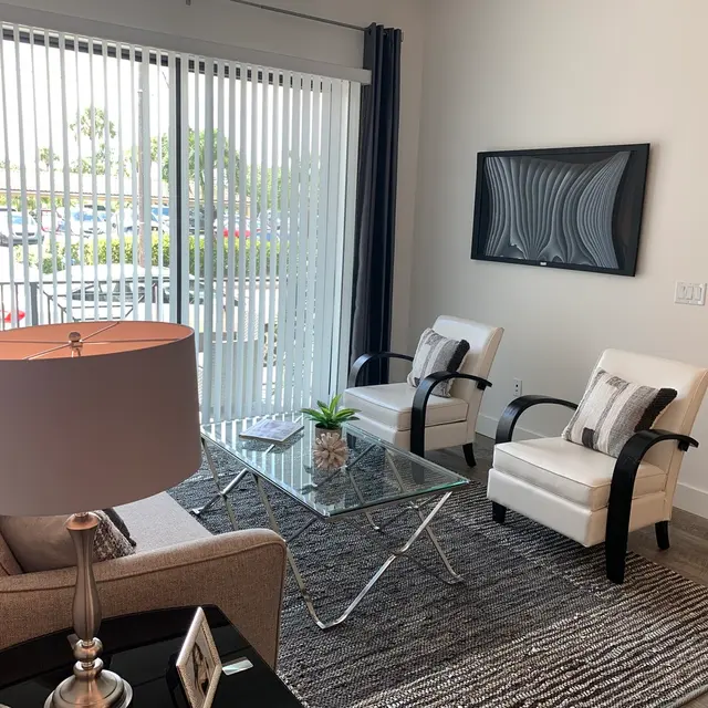 Modern Living Room Design A modern living room with a light-colored sofa, two white armchairs, a glass coffee table, and a lamp. Large glass doors with vertical blinds provide natural light and a view of the outside.