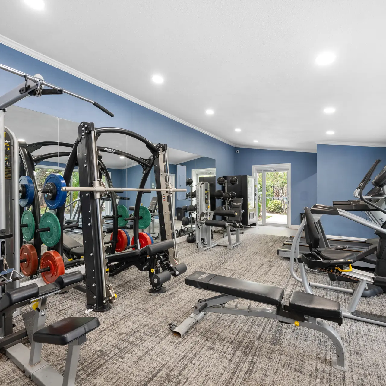 A spacious, modern gym featuring various exercise equipment including weight machines, a multi-gym station with weights, and cardio machines such as an elliptical. Large mirrors line one wall, and there are windows letting in natural light.