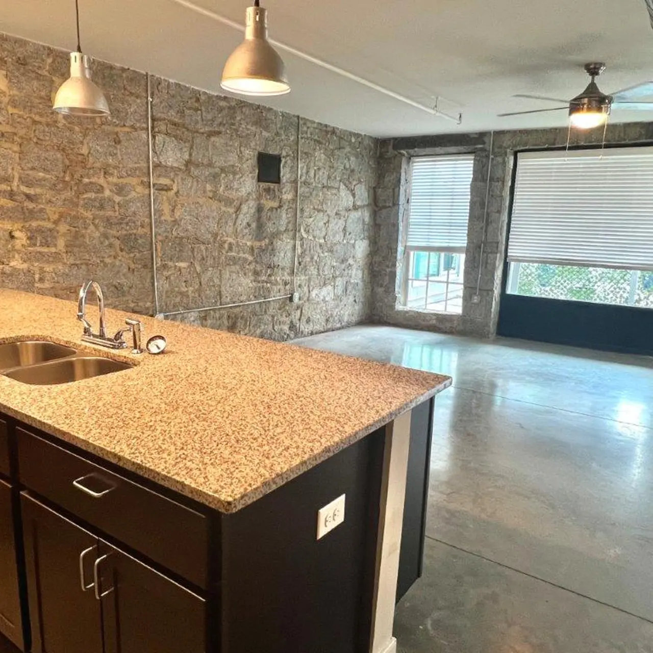 An open-concept living space featuring a kitchen with a granite countertop and dark cabinets, adjacent to a living area with industrial stone walls and large windows.