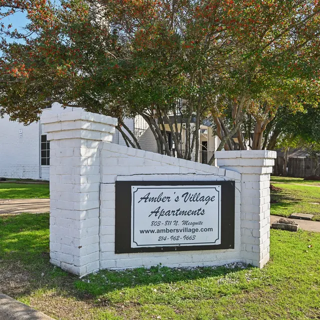 Sign for Amber's Village Apartments situated in a green area with trees and sidewalk.