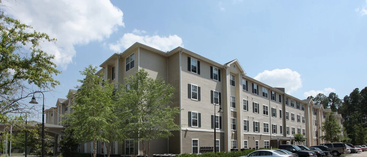 A modern three-story building with multiple windows, surrounded by greenery and parked cars.