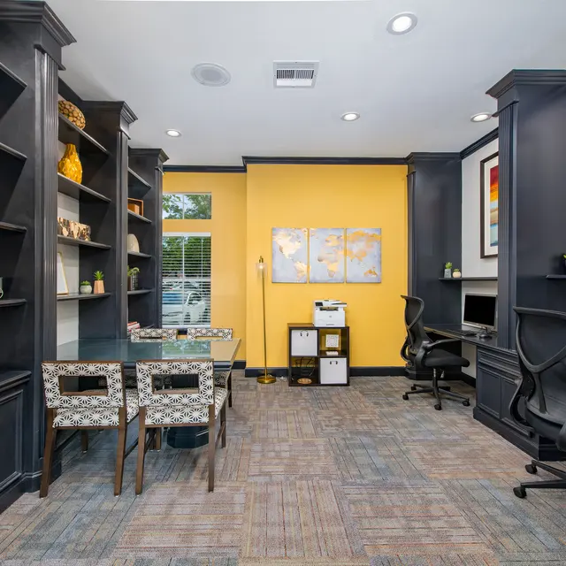 A stylish office space featuring dark wood shelves filled with decorative items and books, a yellow wall, and two desks with computers. There are geometric patterned chairs around a central table.