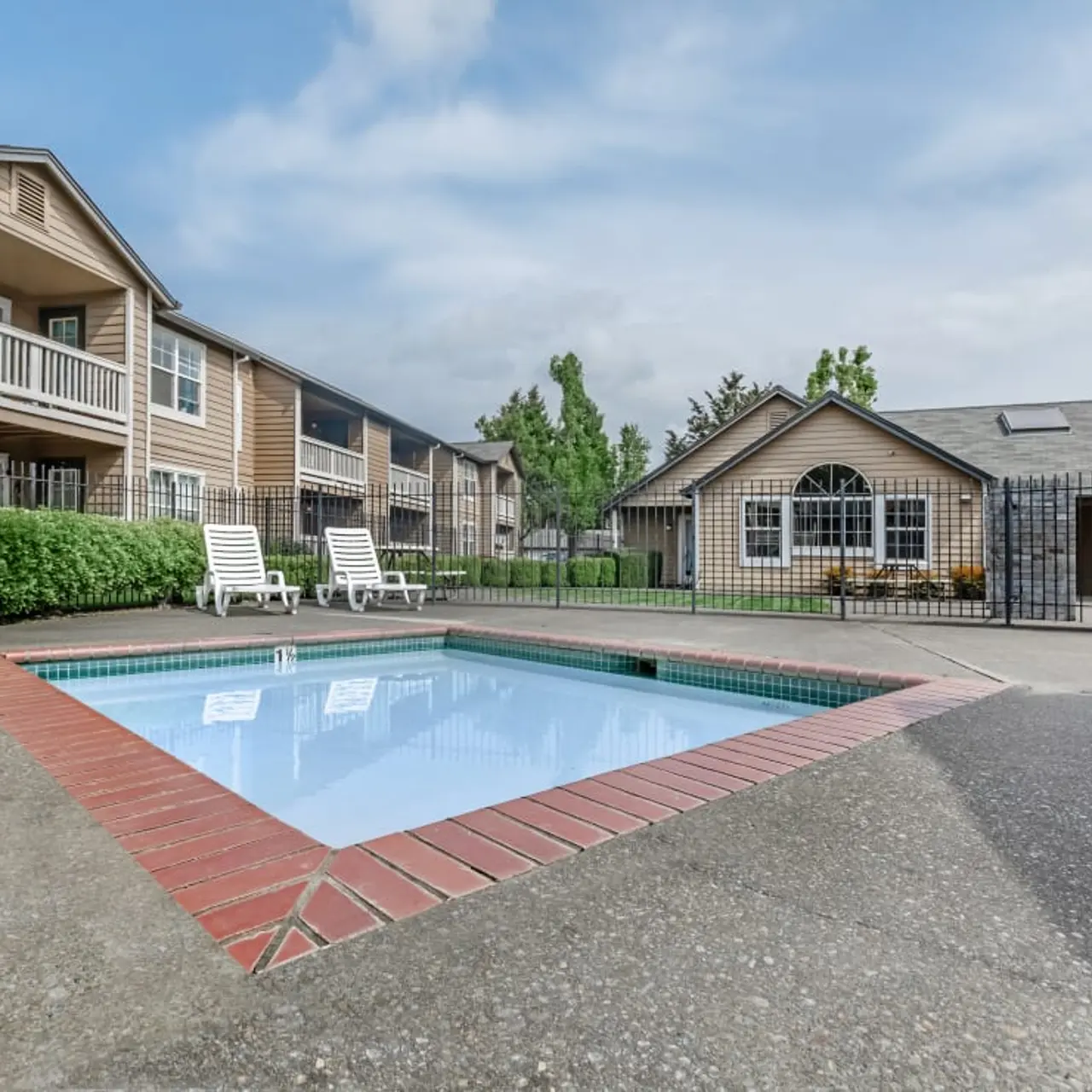 A view of a small swimming pool surrounded by paved area and lounging chairs, with apartment buildings in the background.