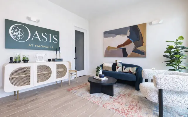 A modern living room featuring a stylish blue couch, a round black coffee table, and a large abstract painting on the wall. The room includes a decorative sideboard and green indoor plants.