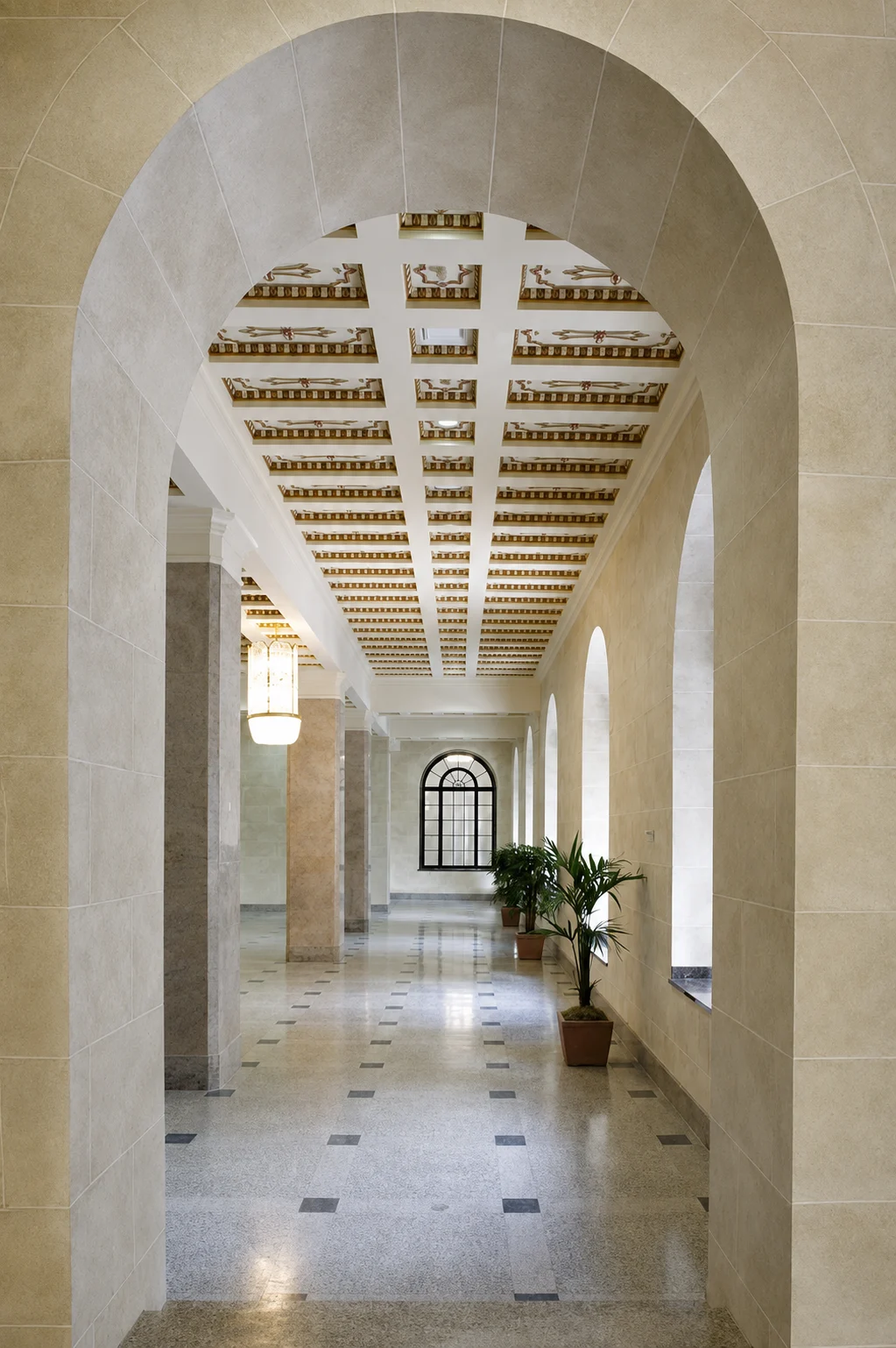A long hallway with arches, columns, and decorative ceiling patterns, featuring potted plants and a large window at the end.