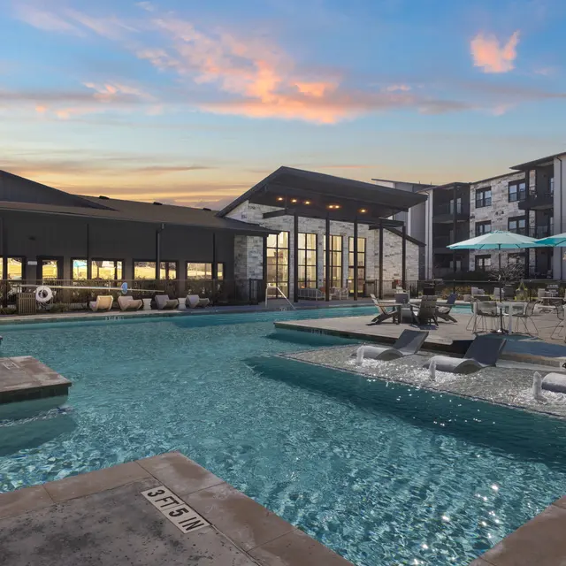 A modern swimming pool area with lounge chairs, umbrellas, and a large building in the background. The setting features a calm evening sky with hues of pink and blue.