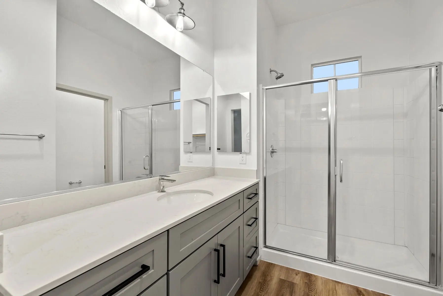 Modern bathroom with a double sink vanity, large mirror, and a glass shower enclosure, featuring light-colored walls and flooring.