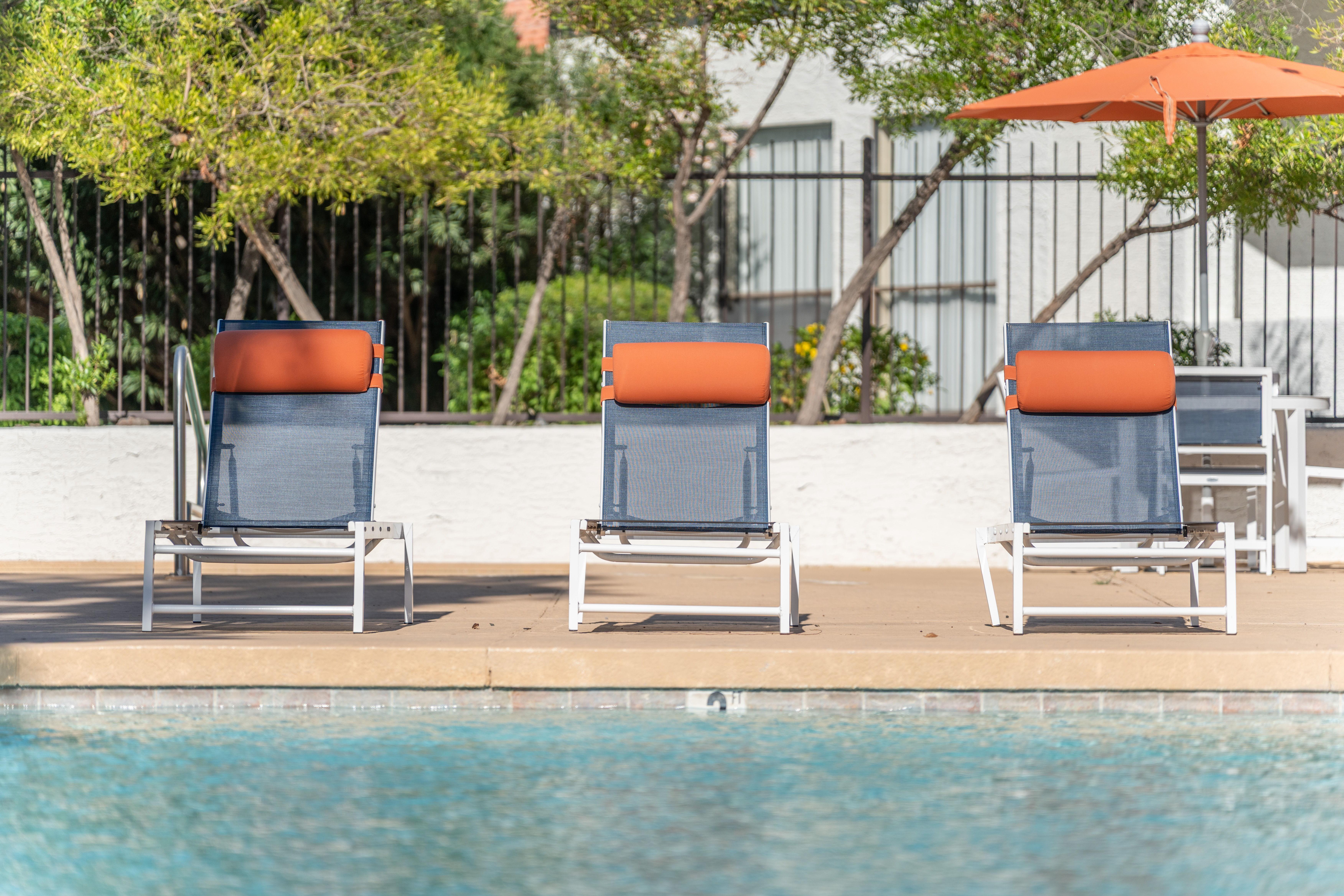 Three loungers by a swimming pool with orange headrests and an orange umbrella nearby.