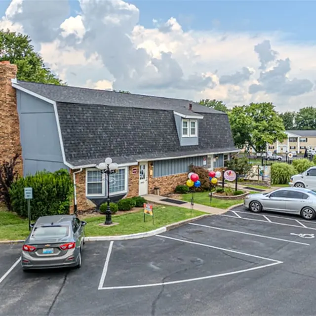A well-landscaped apartment complex with a stone chimney, surrounded by a parking lot filled with cars. Colorful balloons are placed near the entrance.