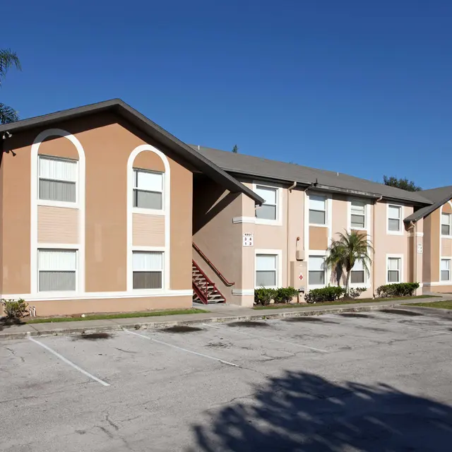 A two-story apartment building with a beige exterior and several windows, surrounded by green landscaping and palm trees. There are several parking spaces in front of the building.