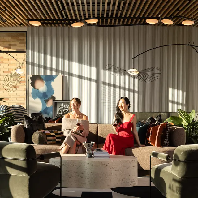 A modern living space featuring two women seated on a plush sofa with stylish decor, plants, and large windows letting in sunlight.