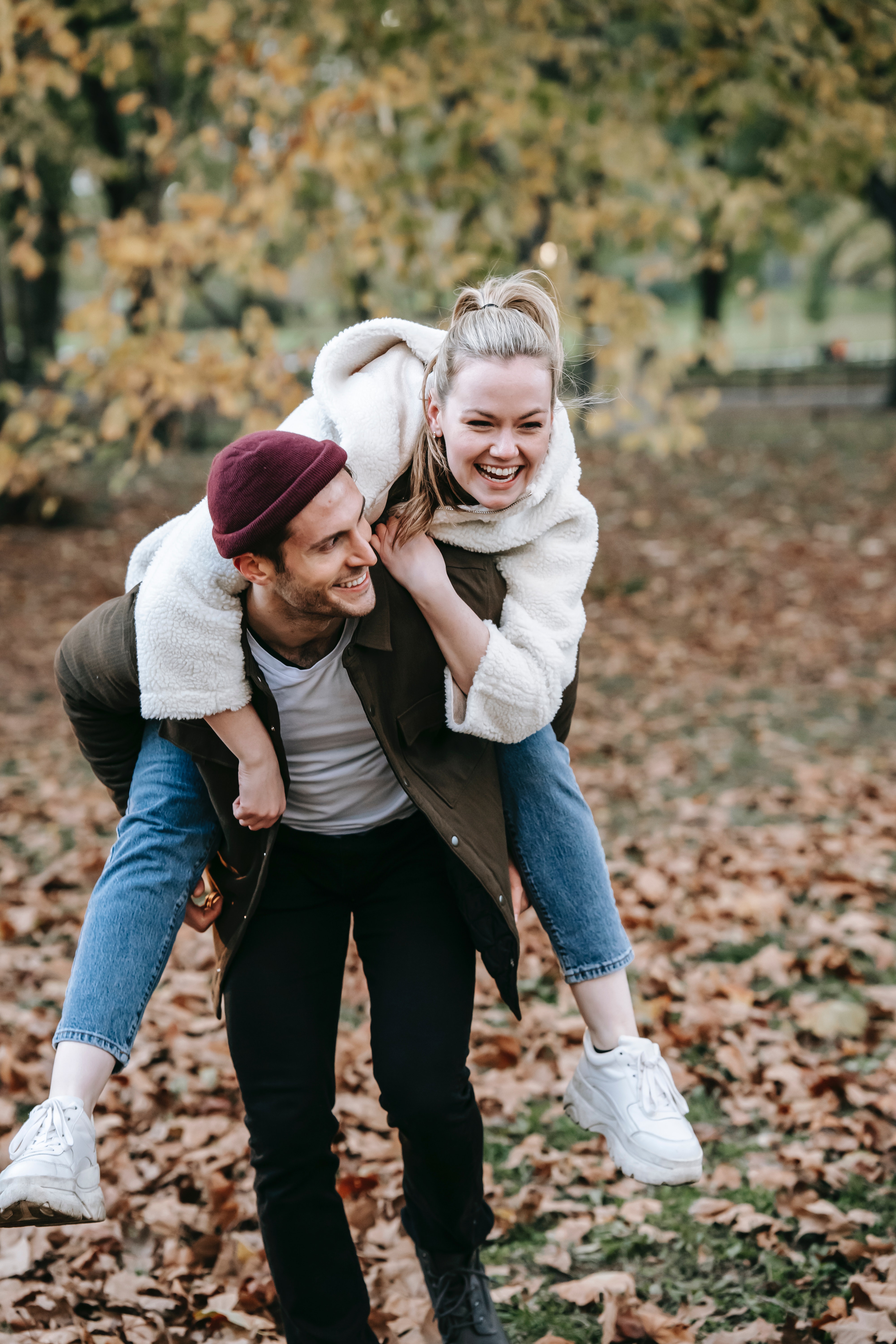 Playful Couple in Autumn Park A young couple enjoying a playful moment in a park during autumn, with fallen leaves around them. The woman is laughing while being carried on the man’s back, both are dressed in casual fall attire.