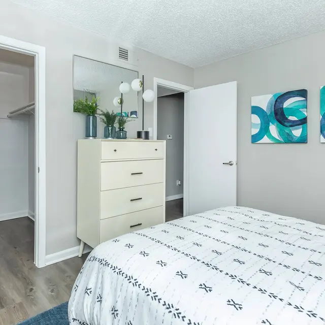 A modern bedroom featuring a white bedspread with black patterns, a light-colored dresser, and abstract wall art in green and blue tones. The room has two doors, one leading to a closet and the other to an exit.