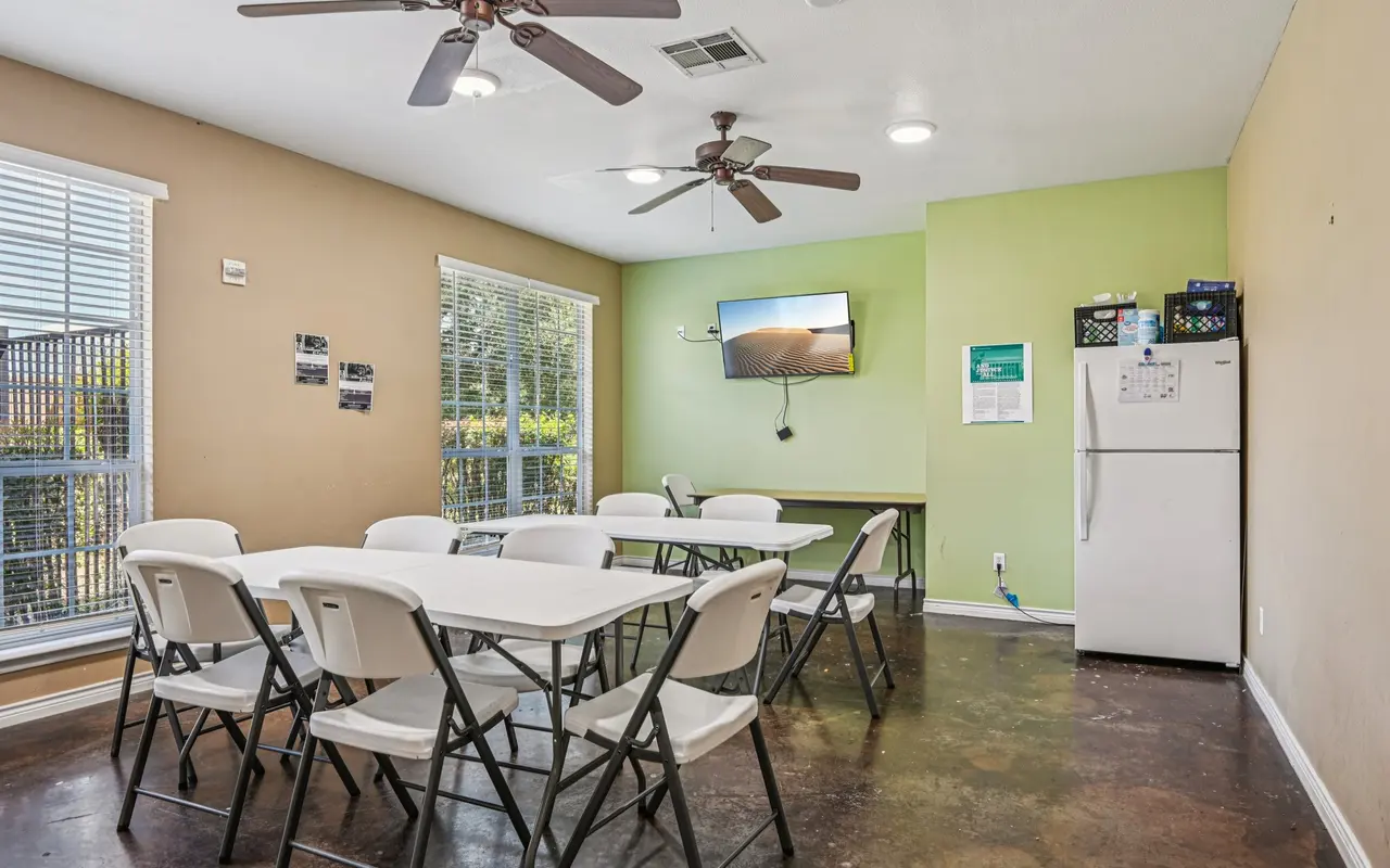 A bright community meeting room featuring a large table with folding chairs, a refrigerator, a TV mounted on the green wall, and windows providing natural light.