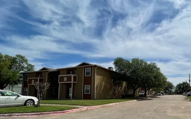 A view of a two-story apartment complex with a grassy area in front and a clear blue sky overhead. Several trees are in the vicinity, and there's a car parked nearby on the street.