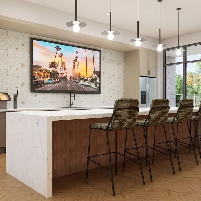 A modern kitchen bar area featuring a large marble countertop, high stools, and a mounted television displaying a street view with palm trees. The space has a stylish design with hanging lights and large windows allowing natural light.