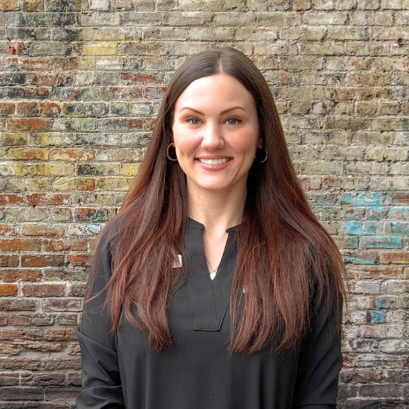 A woman with long brown hair smiles in front of a textured brick wall. She is wearing a black top and has earrings.