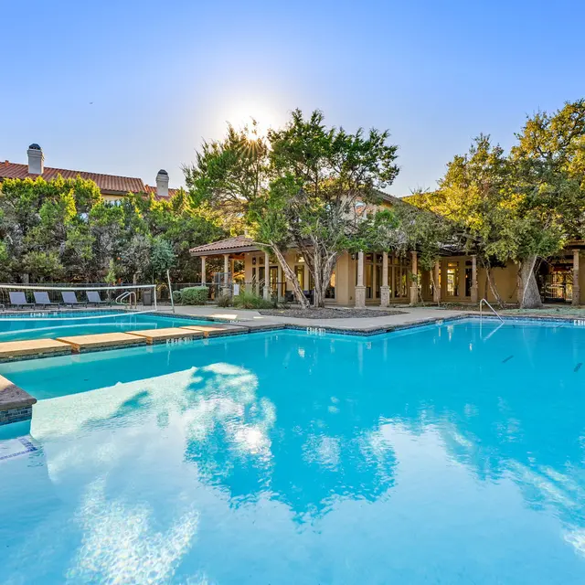 A serene swimming pool view in a resort with clear blue water, surrounded by trees and lounge chairs, set against a clear sky and warm sunlight.