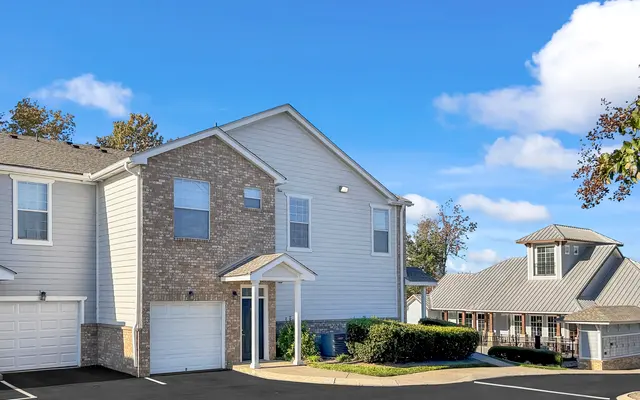 A modern two-story apartment building made of beige and gray siding, with a small garden in front. The sky is bright blue with a few clouds.