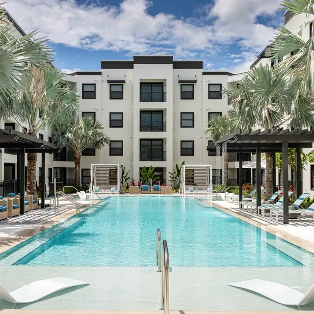 A modern pool area surrounded by palm trees and contemporary architecture. The pool is clear and inviting, with lounge chairs on the side and light clouds in a blue sky above.