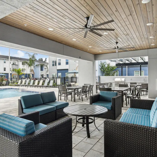 Outdoor lounge area with comfortable seating, a ceiling fan, and a view of a pool surrounded by modern buildings.