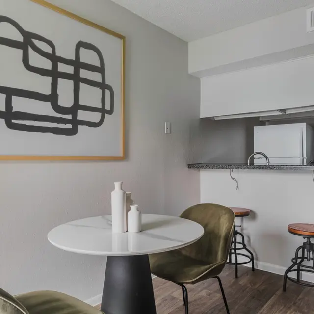 A modern kitchenette area featuring a round white table with two chairs, decorated with minimalist bottles. In the background, there’s a small kitchen with a metal countertop and a white refrigerator, alongside a large abstract art piece on the wall.