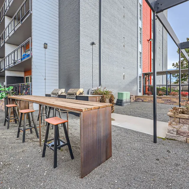 An outdoor space featuring a long wooden table with high stools, surrounded by gravel and a brick border. In the background, there are multiple floors of an apartment building. Part of the scene includes green plants and a shaded area with structures.
