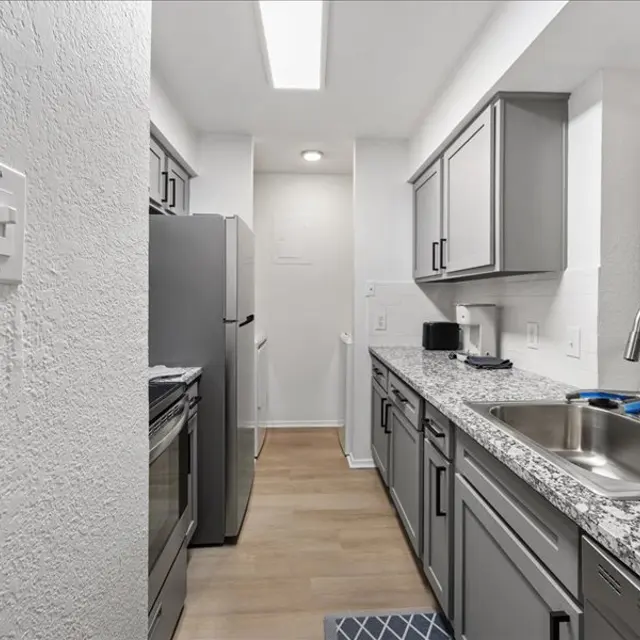 A modern kitchen with gray cabinets, stainless steel appliances, and a granite countertop. The space has a bright and clean design with a sink and some kitchen accessories visible.