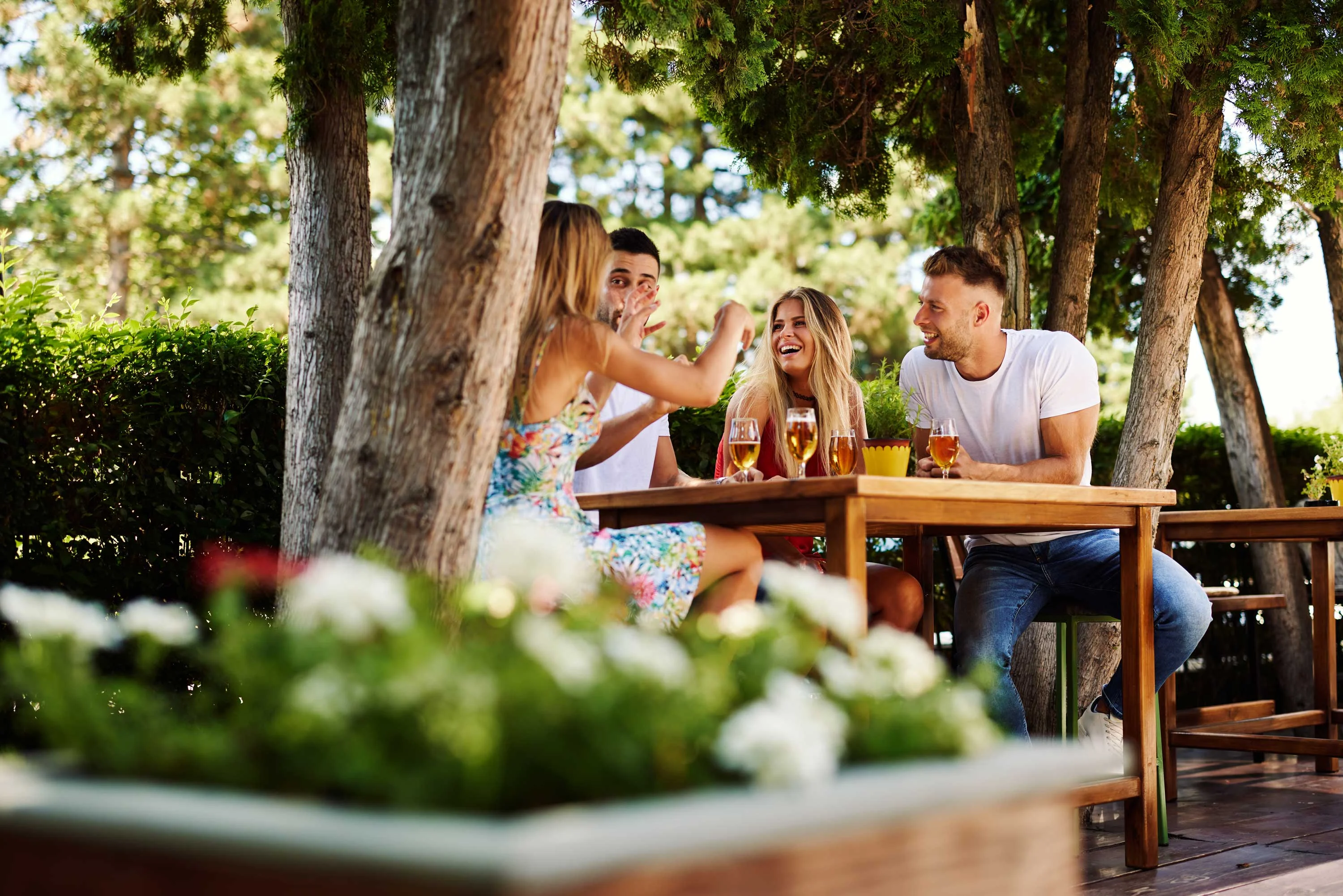A group of four friends enjoying drinks at an outdoor table surrounded by greenery.