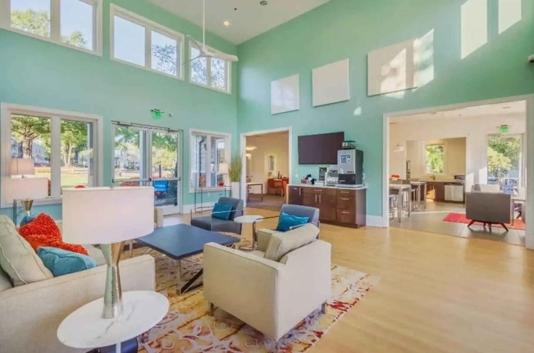 A modern community lounge with bright blue walls, large windows, and wooden flooring. The space features comfortable seating with cream and turquoise furniture, a coffee table, and a television. The open kitchen area is visible in the background, and the room is filled with natural light from the large windows.