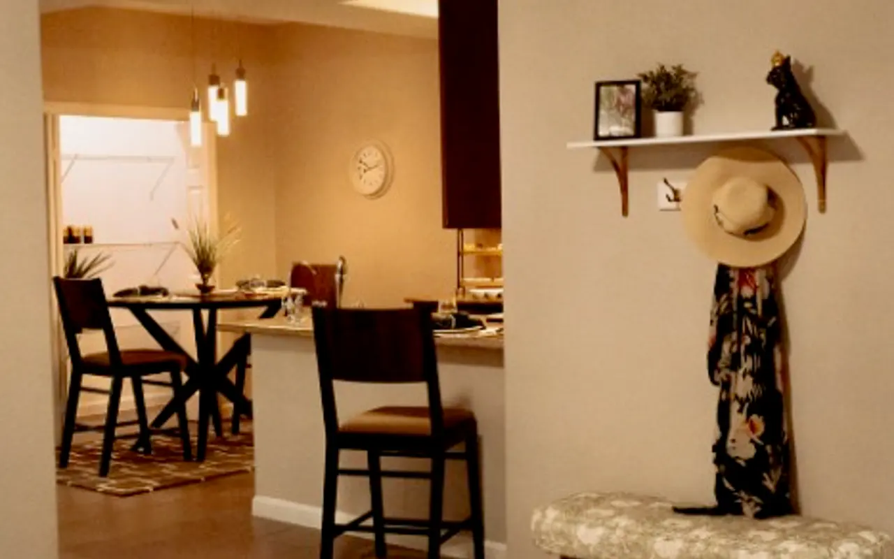 Cozy Living Room Overview A cozy living room with a beige couch and a decorative chair, featuring a ceiling fan and a small shelf with decor. An open doorway leads to a dining area with a table and chairs in the background.